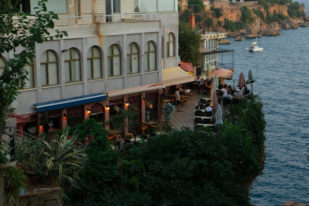An outdoor restaurant terrace is nestled beside a building, overlooking a scenic coastline. Lush green plants surround the area, enhancing the serene ambiance. Several tables with umbrellas are arranged on the terrace, where people are dining and enjoying the seaside view. In the background, there is a calm body of water with a small boat, against a backdrop of cliffs and rustic buildings.