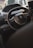 Interior view of a car focusing on the steering wheel with the Peugeot logo. The dashboard and part of the infotainment system are visible, with soft lighting suggesting a late afternoon or evening setting. Residential buildings are blurred in the background through the windshield.