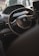 Interior view of a car focusing on the steering wheel with the Peugeot logo. The dashboard and part of the infotainment system are visible, with soft lighting suggesting a late afternoon or evening setting. Residential buildings are blurred in the background through the windshield.