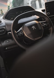 Interior view of a car focusing on the steering wheel with the Peugeot logo. The dashboard and part of the infotainment system are visible, with soft lighting suggesting a late afternoon or evening setting. Residential buildings are blurred in the background through the windshield.
