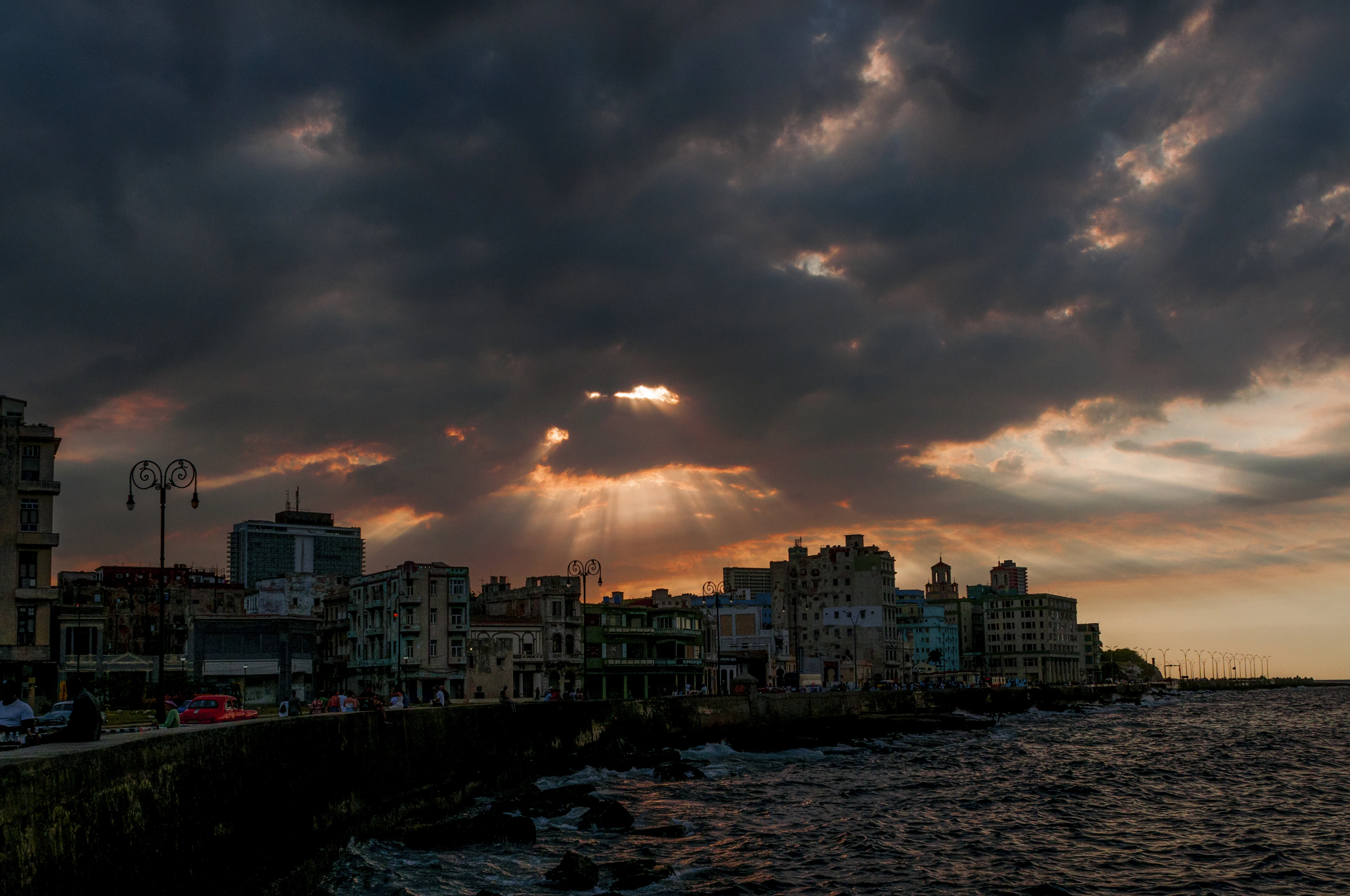 Sunlight breaking through heavy cloud over Havana's Malecon at dusk, the sea wall curving past a long terrace of weathered apartment blocks while waves break on the rocks below.