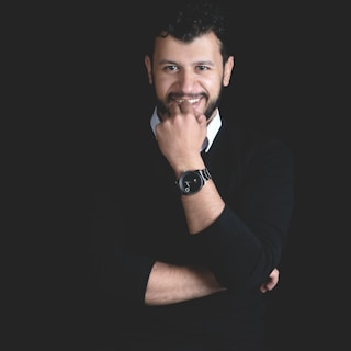A man with dark curly hair and a beard stands against a dark background. He is wearing a black sweater over a collared shirt and a watch on his wrist, with one hand touching his chin and the other arm crossed.