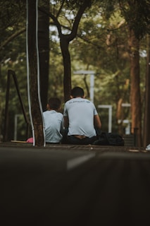 Two individuals are sitting on a wooden platform in a park-like setting, surrounded by trees and greenery. They are both facing away, and one of them is wearing a shirt with a logo visible. The atmosphere is calm and secluded, with dappled sunlight filtering through the leaves.