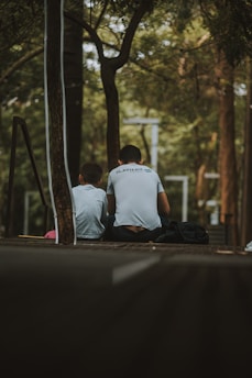 Two individuals are sitting on a wooden platform in a park-like setting, surrounded by trees and greenery. They are both facing away, and one of them is wearing a shirt with a logo visible. The atmosphere is calm and secluded, with dappled sunlight filtering through the leaves.