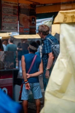 A young person wearing a blue sleeveless shirt, blue shorts, and colorful wristbands stands with their hands clasped behind their back, next to an older person in a plaid shirt and glasses. They appear to be observing a stall with signs advertising various food items in Portuguese. The atmosphere resembles an outdoor market or fair.