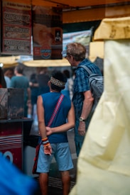 A young person wearing a blue sleeveless shirt, blue shorts, and colorful wristbands stands with their hands clasped behind their back, next to an older person in a plaid shirt and glasses. They appear to be observing a stall with signs advertising various food items in Portuguese. The atmosphere resembles an outdoor market or fair.