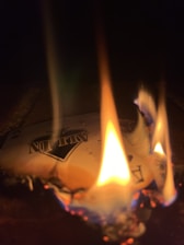 A playing card is on fire, with flames consuming its edges. The card displays the word 'ALL IN' and features a spade symbol. The scene is dimly lit, emphasizing the bright and colorful flames.