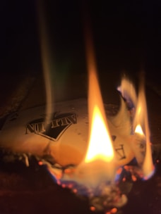 A playing card is on fire, with flames consuming its edges. The card displays the word 'ALL IN' and features a spade symbol. The scene is dimly lit, emphasizing the bright and colorful flames.
