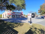A fast-food restaurant building with a combination of KFC and Taco Bell logos on the facade, featuring red and white striped awnings. The restaurant is set in a parking lot surrounded by trees with a clear blue sky overhead. An exit sign in the foreground includes both KFC and Taco Bell logos.