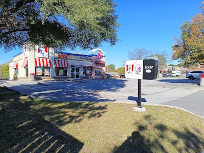 A fast-food restaurant building with a combination of KFC and Taco Bell logos on the facade, featuring red and white striped awnings. The restaurant is set in a parking lot surrounded by trees with a clear blue sky overhead. An exit sign in the foreground includes both KFC and Taco Bell logos.