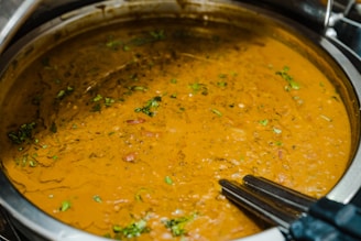 A pot filled with a thick, brown lentil soup or dhal, garnished with fresh green herbs. The surface has a slightly textured look with visible chunks of vegetables and spices.