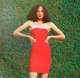 A person with curly hair is wearing a bright red strapless dress and posing confidently with hands on hips against a lush green leafy background.