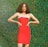 A person with curly hair is wearing a bright red strapless dress and posing confidently with hands on hips against a lush green leafy background.