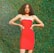 A person with curly hair is wearing a bright red strapless dress and posing confidently with hands on hips against a lush green leafy background.