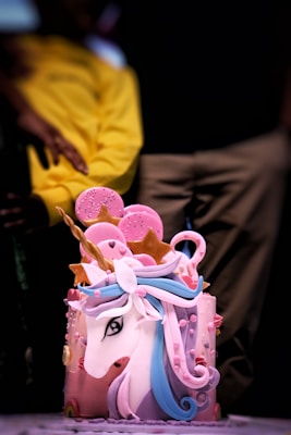 A whimsical cake designed to look like a unicorn, featuring a white face with a colorful mane in blue and pink tones. The cake is adorned with star and circle decorations, primarily in pink, and a golden horn. It appears to be placed on a plain background with a person in yellow clothing partially visible in the blurred background.