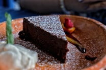 A rich, dark chocolate cake slice garnished with powdered sugar sits on a rustic wooden plate. Next to it, a dollop of whipped cream is topped with a green garnish. In the background, vibrant red raspberries and orange segments add a pop of color.