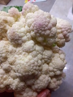 A close-up view of a cauliflower head with tightly packed florets. The texture is bumpy and uneven, with hints of purple in some areas. The background includes a tabletop surface, adding context to the setting.