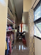 A narrow, well-organized closet with shelves on the left housing folded clothes and clothes on hangers. The closet has a glossy floor reflecting light from a small window at the end. An orange curtain hangs at the window, complementing the yellow and brown patterned curtain on the right. There are bags and clothing items on a small table beneath the window.