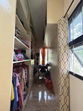 A narrow, well-organized closet with shelves on the left housing folded clothes and clothes on hangers. The closet has a glossy floor reflecting light from a small window at the end. An orange curtain hangs at the window, complementing the yellow and brown patterned curtain on the right. There are bags and clothing items on a small table beneath the window.