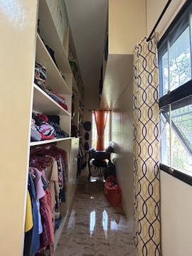 A narrow, well-organized closet with shelves on the left housing folded clothes and clothes on hangers. The closet has a glossy floor reflecting light from a small window at the end. An orange curtain hangs at the window, complementing the yellow and brown patterned curtain on the right. There are bags and clothing items on a small table beneath the window.