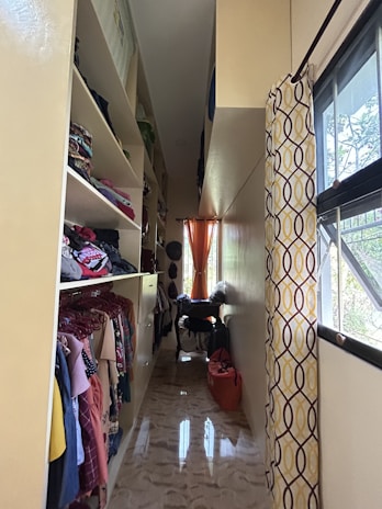A narrow, well-organized closet with shelves on the left housing folded clothes and clothes on hangers. The closet has a glossy floor reflecting light from a small window at the end. An orange curtain hangs at the window, complementing the yellow and brown patterned curtain on the right. There are bags and clothing items on a small table beneath the window.