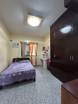 A neat and orderly bedroom with a single bed covered with a purple bedspread on the left side, next to a window with two sets of curtains. Below the window is an air conditioning unit. To the right of the bed is a desk with a white chair, a lamp, and some stuffed animals on top of a shelving unit. On the far right, there is a large, dark-colored wardrobe that takes up most of the wall. The room has beige walls and a patterned tiled floor. A large ceiling light fixture is centered on the ceiling.