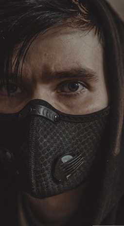 A close-up of a person wearing a dark, hexagonal patterned face mask with vent filters, partially covered by a hood. The person has intense eyes and dark hair.