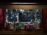 A tightly packed shoe store with walls and shelves filled with various types of footwear including sandals, sneakers, and boots. The shoes are organized by style and color, with numerous shoe boxes stacked on the floor. A person is seen inside the store, possibly attending to the shop.