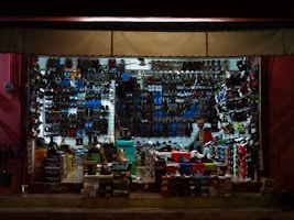 A tightly packed shoe store with walls and shelves filled with various types of footwear including sandals, sneakers, and boots. The shoes are organized by style and color, with numerous shoe boxes stacked on the floor. A person is seen inside the store, possibly attending to the shop.