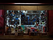 A tightly packed shoe store with walls and shelves filled with various types of footwear including sandals, sneakers, and boots. The shoes are organized by style and color, with numerous shoe boxes stacked on the floor. A person is seen inside the store, possibly attending to the shop.