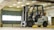 A forklift is situated in an indoor garage or warehouse environment. It is a Nissan model with a large lift mechanism and the designation 'B38' on its side. The area is well-lit with fluorescent lights, and a large industrial-style window is visible in the background along with concrete beams and walls.