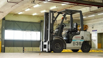 A forklift is situated in an indoor garage or warehouse environment. It is a Nissan model with a large lift mechanism and the designation 'B38' on its side. The area is well-lit with fluorescent lights, and a large industrial-style window is visible in the background along with concrete beams and walls.