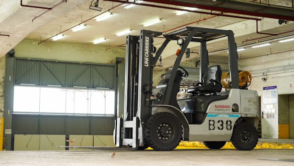 A forklift is situated in an indoor garage or warehouse environment. It is a Nissan model with a large lift mechanism and the designation 'B38' on its side. The area is well-lit with fluorescent lights, and a large industrial-style window is visible in the background along with concrete beams and walls.