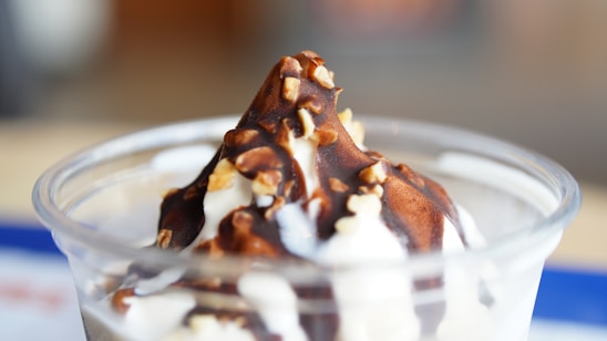 A creamy vanilla ice cream topped with a rich chocolate sauce and sprinkled with crushed nuts, served in a clear cup. The background is softly blurred, emphasizing the smooth texture and vibrant colors of the dessert.