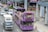 A purple double-decker sightseeing bus filled with tourists travels through an urban area. Many people on the top deck are wearing hats, and some are taking photos. The bus displays digital screens with messages such as 'WELCOME T'. Nearby, cars and a truck are sharing the road. The surroundings include modern buildings and a sidewalk with pedestrians.