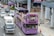 A purple double-decker sightseeing bus filled with tourists travels through an urban area. Many people on the top deck are wearing hats, and some are taking photos. The bus displays digital screens with messages such as 'WELCOME T'. Nearby, cars and a truck are sharing the road. The surroundings include modern buildings and a sidewalk with pedestrians.