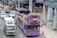 A purple double-decker sightseeing bus filled with tourists travels through an urban area. Many people on the top deck are wearing hats, and some are taking photos. The bus displays digital screens with messages such as 'WELCOME T'. Nearby, cars and a truck are sharing the road. The surroundings include modern buildings and a sidewalk with pedestrians.