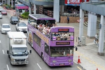 A purple double-decker sightseeing bus filled with tourists travels through an urban area. Many people on the top deck are wearing hats, and some are taking photos. The bus displays digital screens with messages such as 'WELCOME T'. Nearby, cars and a truck are sharing the road. The surroundings include modern buildings and a sidewalk with pedestrians.