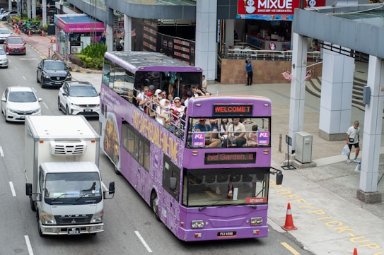 A purple double-decker sightseeing bus filled with tourists travels through an urban area. Many people on the top deck are wearing hats, and some are taking photos. The bus displays digital screens with messages such as 'WELCOME T'. Nearby, cars and a truck are sharing the road. The surroundings include modern buildings and a sidewalk with pedestrians.