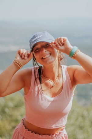A woman wearing a blue cap and a pink sleeveless top smiles brightly while holding the brim of her cap with both hands. She accessorizes with golden bracelets and a teal wristband, and stands outdoors with a blurred natural background.