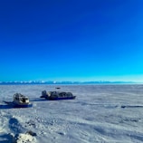 An expansive icy landscape stretches out under a vivid blue sky with snow-covered mountains in the distance. Two vehicles designed for snow travel are parked on the icy terrain, highlighting the remote and isolated nature of the setting.