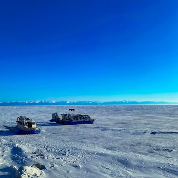 An expansive icy landscape stretches out under a vivid blue sky with snow-covered mountains in the distance. Two vehicles designed for snow travel are parked on the icy terrain, highlighting the remote and isolated nature of the setting.