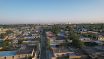 An aerial view of a residential area with small houses arranged in a grid and surrounded by trees. The houses have flat roofs, some of which are painted blue. A long, straight road runs through the center of the image, leading towards the horizon with more densely packed buildings visible in the distance.