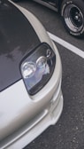 A close-up view of the front section of a sleek car, showcasing a modern headlight design with a glossy finish. The car features a carbon fiber hood and a silver body, parked on a textured asphalt surface with another vehicle in the background.