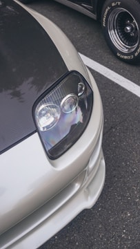 A close-up view of the front section of a sleek car, showcasing a modern headlight design with a glossy finish. The car features a carbon fiber hood and a silver body, parked on a textured asphalt surface with another vehicle in the background.