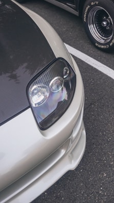 A close-up view of the front section of a sleek car, showcasing a modern headlight design with a glossy finish. The car features a carbon fiber hood and a silver body, parked on a textured asphalt surface with another vehicle in the background.
