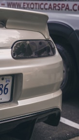 The image captures the rear end of a white sports car, showcasing a spoiler, tail lights, and part of a license plate. In the background, there is a sign that appears to be part of a business advertisement with the text partially visible.