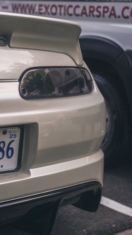 The image captures the rear end of a white sports car, showcasing a spoiler, tail lights, and part of a license plate. In the background, there is a sign that appears to be part of a business advertisement with the text partially visible.