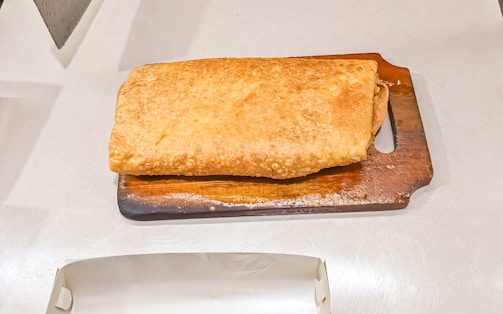 A rectangular, golden-brown pastry is placed on a small wooden cutting board. The pastry has a flaky texture and occupies most of the board. The surface is evenly baked, and its edges are slightly curved. The wooden board has a handle and sits on a light-colored surface.