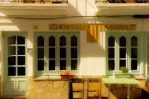 A quaint facade of a small hostal featuring light green doors and windows set in a cream-colored wall. The sign above reads 'Hostal Maria-Rosa'. There are two chairs and a small table with a floral pattern in front, and a potted plant sits on the windowsill.